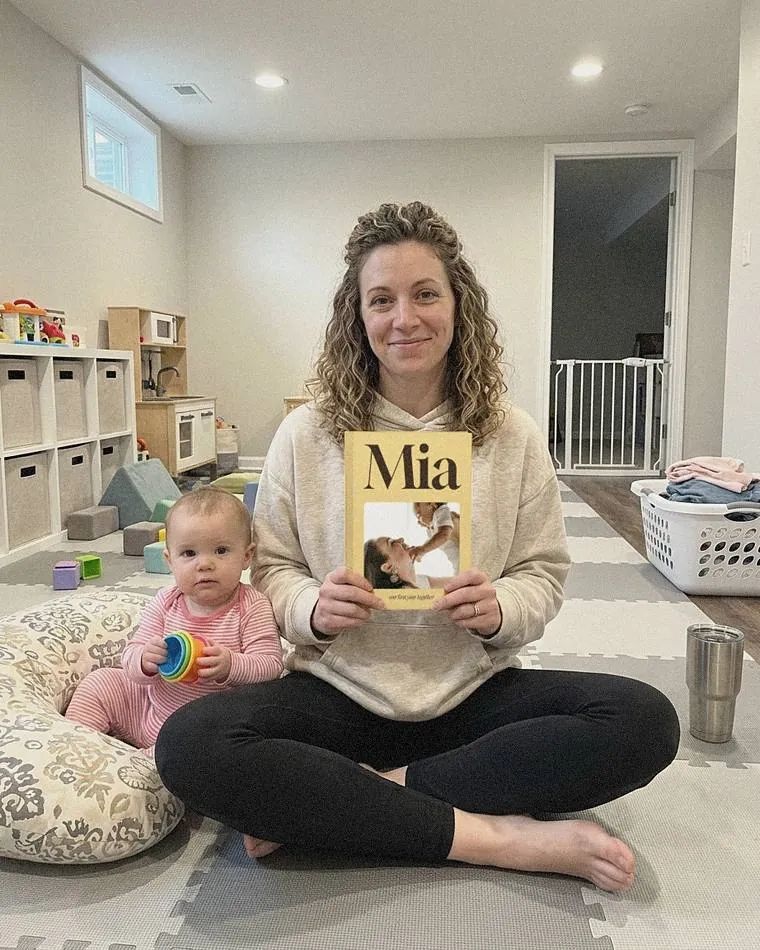 Mom sitting on the playroom floor holding her yellow Mia book, with her baby daughter beside her