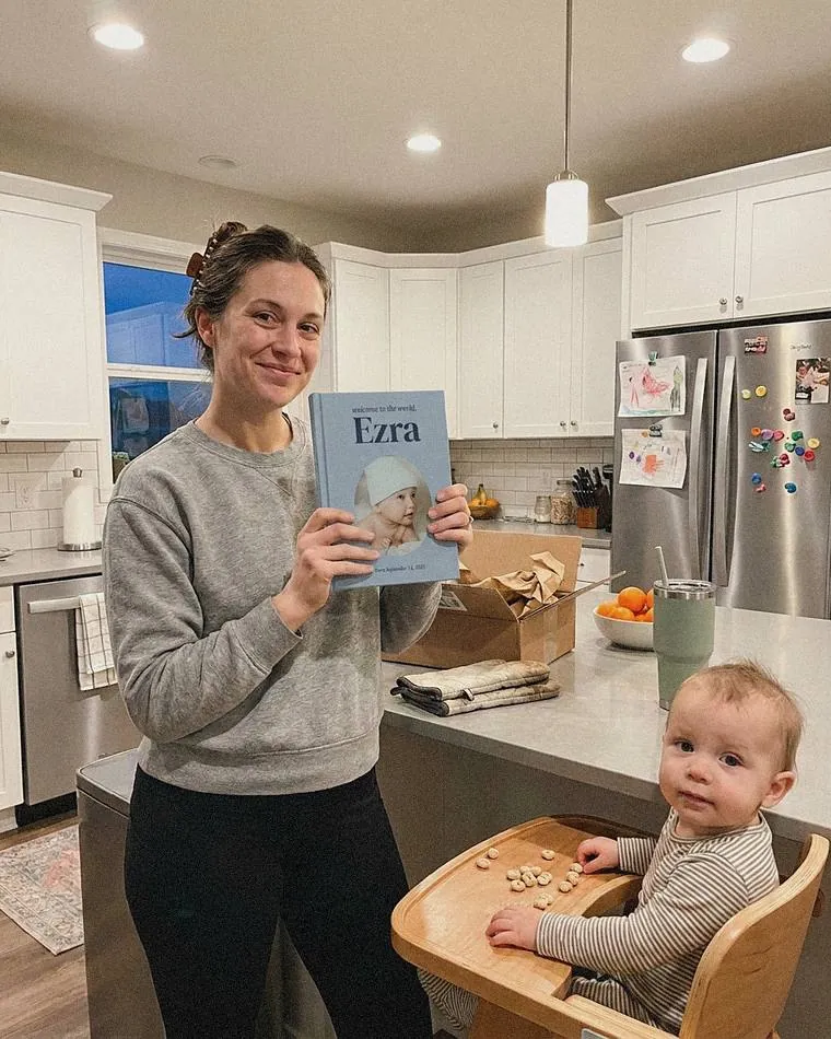 Mom in her kitchen holding her Welcome to the world, Ezra book, with her son in the high chair beside her
